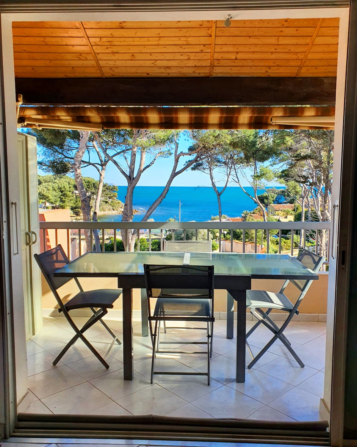 A dining area features a glass-topped table surrounded by four folding chairs, positioned on a balcony. The view of the sea is visible through large sliding glass doors, framed by trees and sunlight filtering through the outdoor awning.