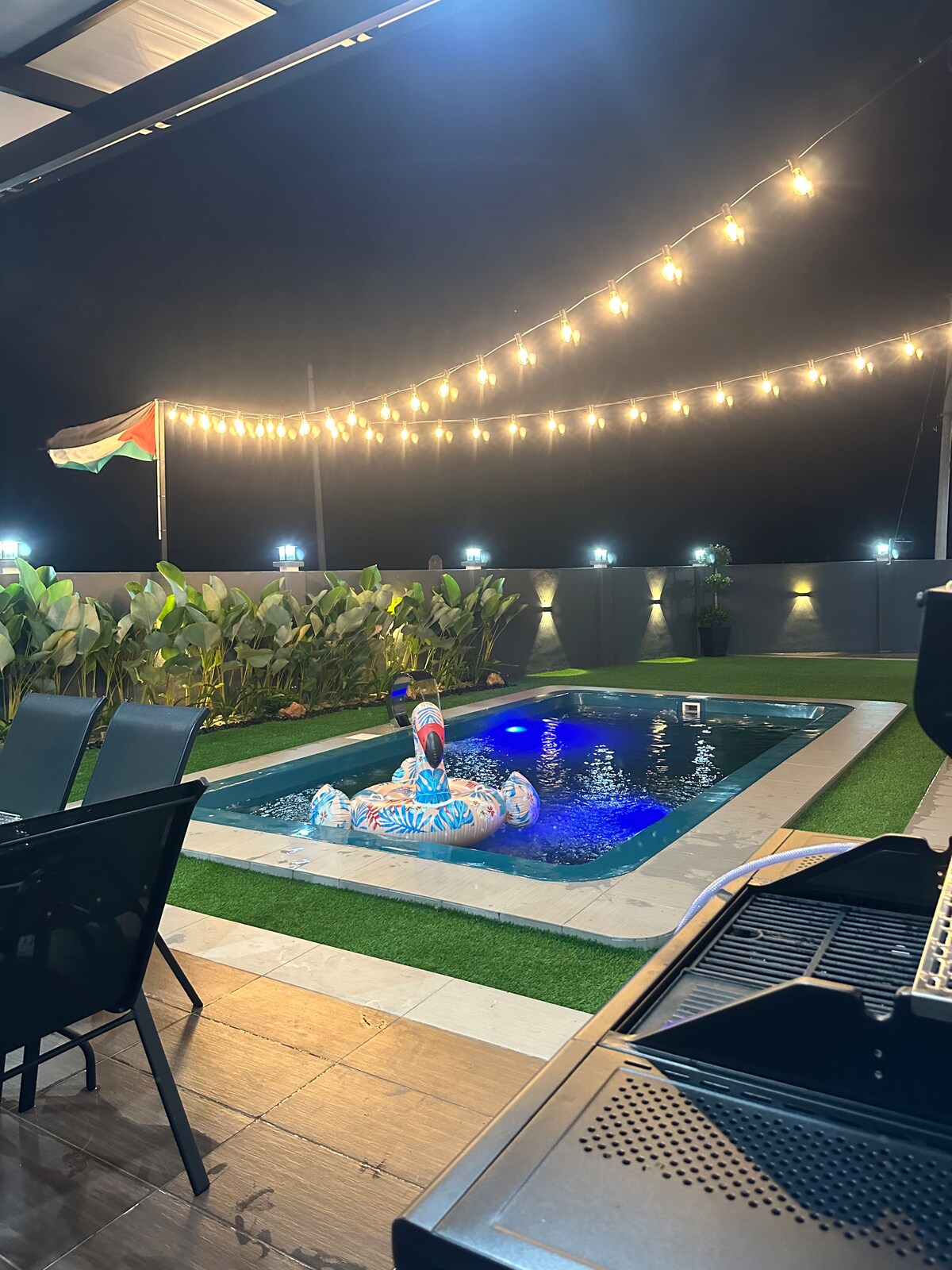 A well-lit outdoor area features a jacuzzi pool surrounded by lush greenery and modern fencing. String lights create a warm ambiance overhead, while a BBQ grill is positioned nearby. Colorful inflatables float on the water's surface, contributing to a relaxed atmosphere.