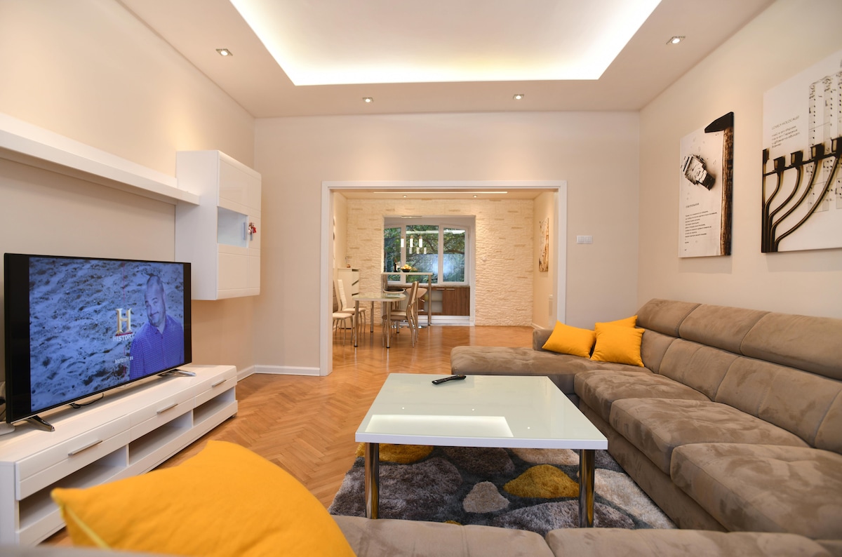 A spacious living area features a comfortable sectional sofa adorned with yellow cushions. A television is positioned on a white stand, facing the seating. The adjacent dining area is visible through an open doorway, showcasing a round table surrounded by chairs.