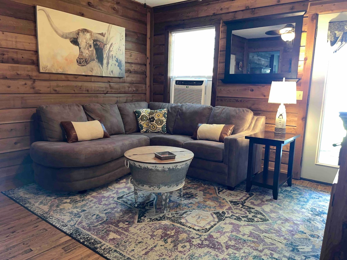 A comfortable seating area features a large sectional sofa adorned with decorative pillows. A rustic coffee table sits atop a colorful area rug. Natural light enters through a window, and a lamp illuminates the space, creating a cozy atmosphere.