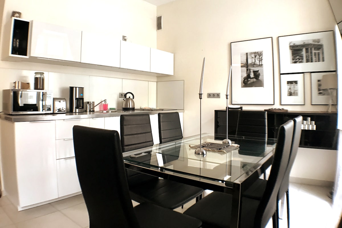 A modern kitchen is shown with sleek white cabinetry and stainless steel appliances. A glass dining table is surrounded by six black chairs. Black and white framed photos adorn the walls, contributing to the contemporary atmosphere.