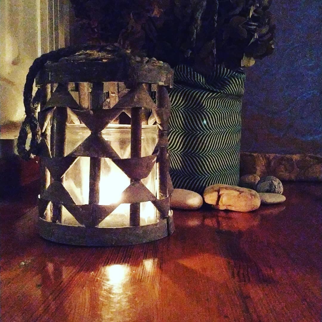 A lantern with a woven design is placed on a wooden surface, gently illuminating its surroundings. Nearby, pebbles are arranged along with a potted plant featuring a patterned pot, enhancing the tranquil atmosphere.