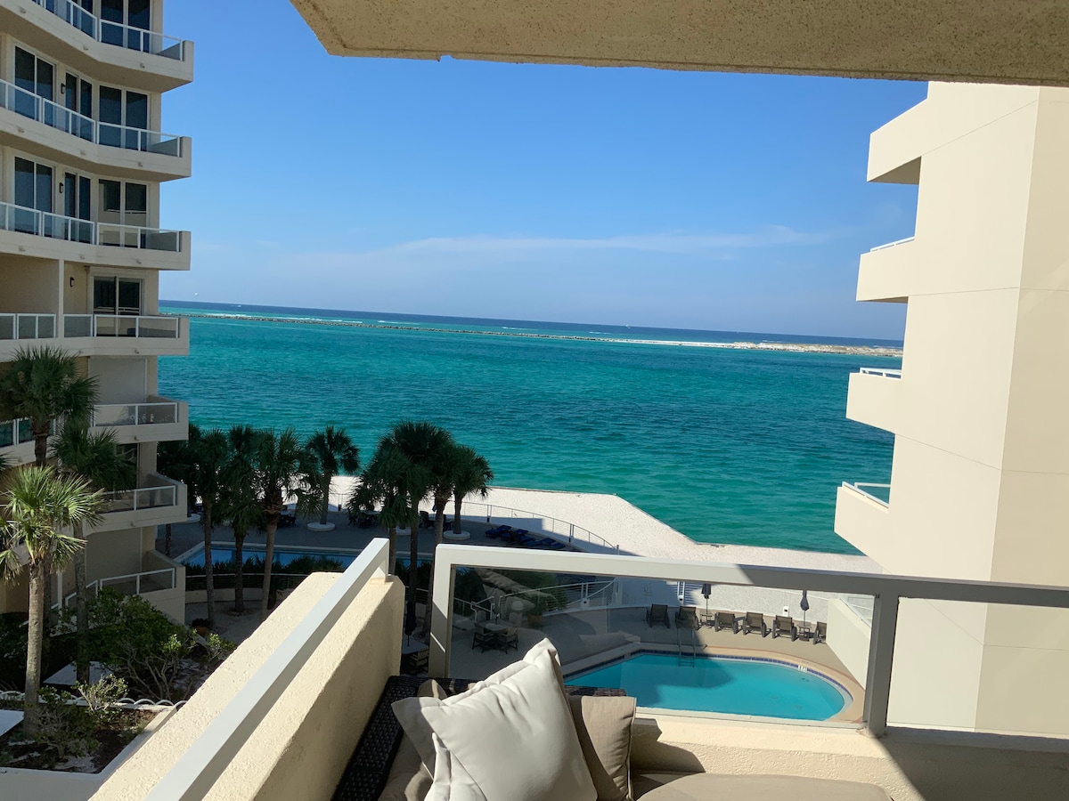 A clear view is offered from the balcony, showcasing tranquil turquoise waters and white sandy beach. Palm trees line the area below, while a pool is visible in the distance. The sky is bright with minimal clouds, enhancing the coastal scenery.