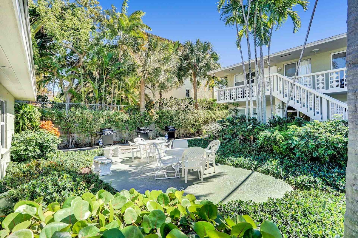A landscaped outdoor area is surrounded by lush greenery and palm trees. A seating arrangement with white furniture is positioned on a patio, complemented by a barbecue grill. The peaceful environment invites relaxation and outdoor dining.