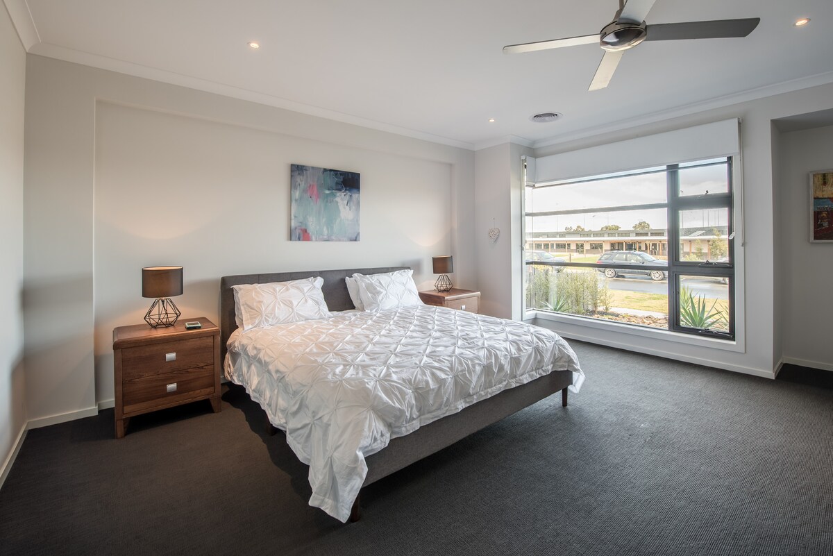 A spacious bedroom features a comfortable bed dressed in white bedding. Two bedside tables hold lamps, providing soft lighting. Large windows offer natural light and a view of the outdoor landscape, while neutral tones create a calm and relaxing environment.