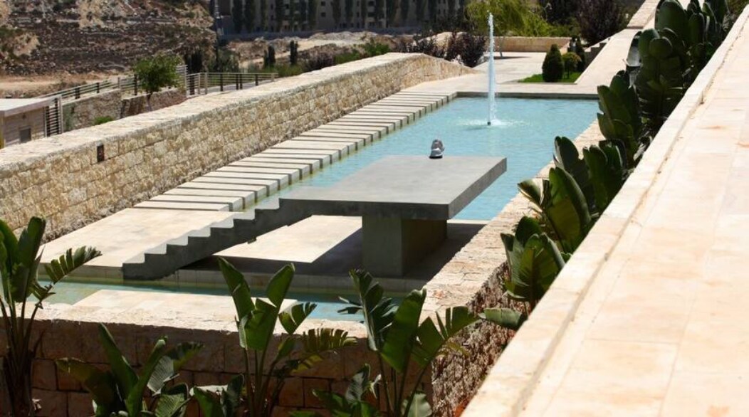 A modern outdoor area is featured, showcasing a long rectangular pool with a central fountain. Minimalist stone pathways run alongside, framed by lush green plants, creating a serene environment. The surrounding landscape offers a backdrop of natural hills and distant buildings.