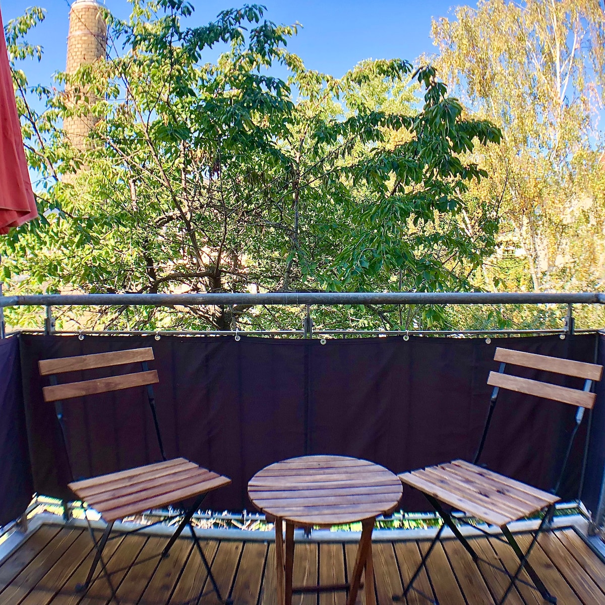 A small balcony is featured with two wooden folding chairs and a round wooden table. Green foliage from surrounding trees provides a natural view, creating a serene outdoor space for relaxation.