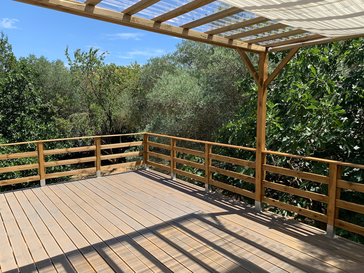 A spacious wooden terrace is surrounded by a lush canopy of greenery, featuring a sturdy railing and an overhead pergola that provides shade. Natural light filters through, creating a calm and inviting outdoor space ideal for relaxation.