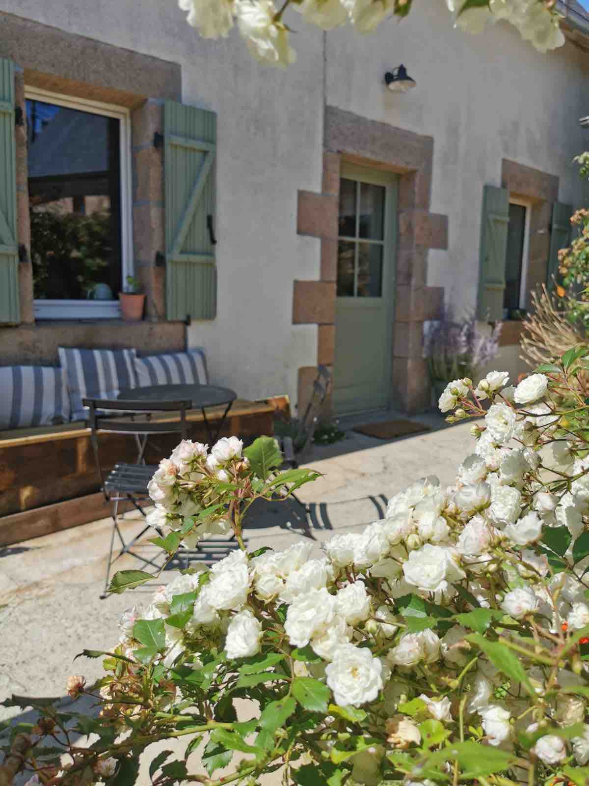 A charming exterior of a home is framed by blooming white roses, with a small seating area featuring a bistro table and chairs. Shuttered windows add a touch of elegance, while a calm and inviting pathway leads to the entrance.