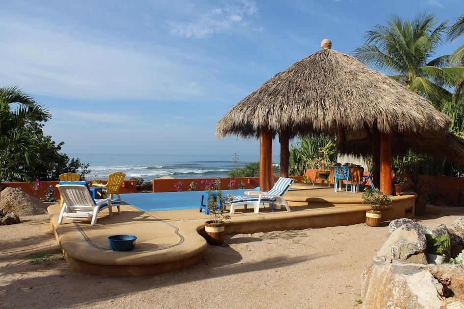 A relaxing outdoor area features a palapa with a thatched roof providing shade over a wooden deck. Lounge chairs and colorful seating are arranged around a pool, with the ocean waves visible in the background. Lush greenery surrounds the space, enhancing the tranquil atmosphere.