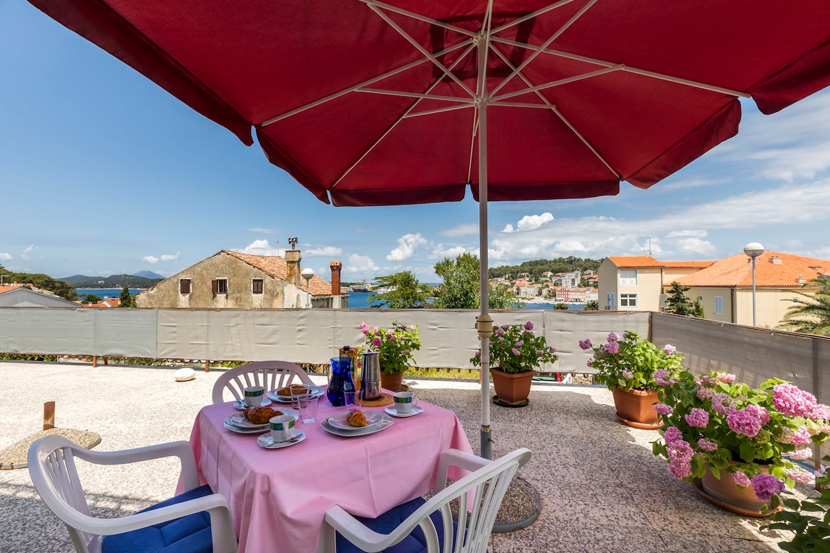 A dining area on the terrace is shaded by a large red umbrella, featuring a table set with plates of food and drink. Surrounding the space are potted plants, including vibrant flowers, with a view of the distant coastline under a clear blue sky.