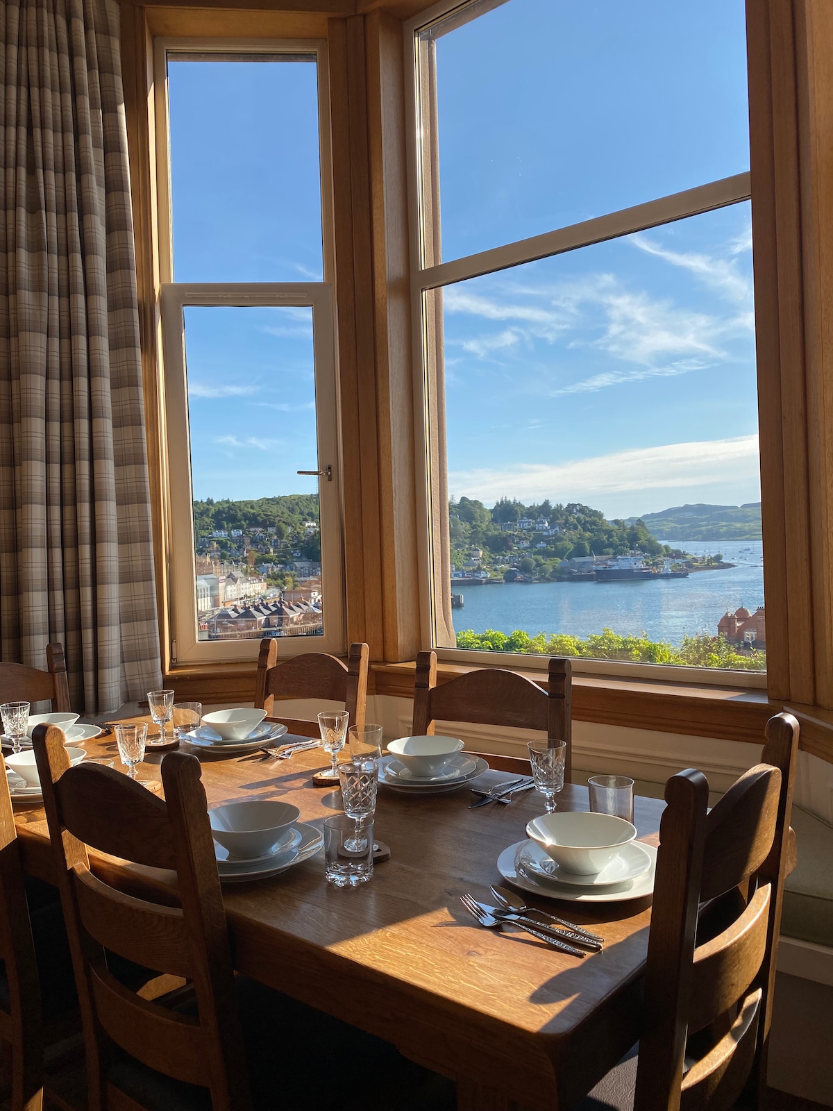 A sunny dining area is anchored by a wooden table set for a meal, featuring plates and glassware. Large windows frame a panoramic view of Oban Bay, allowing natural light to fill the space. The soft fabric curtains complement the warm wood tones.