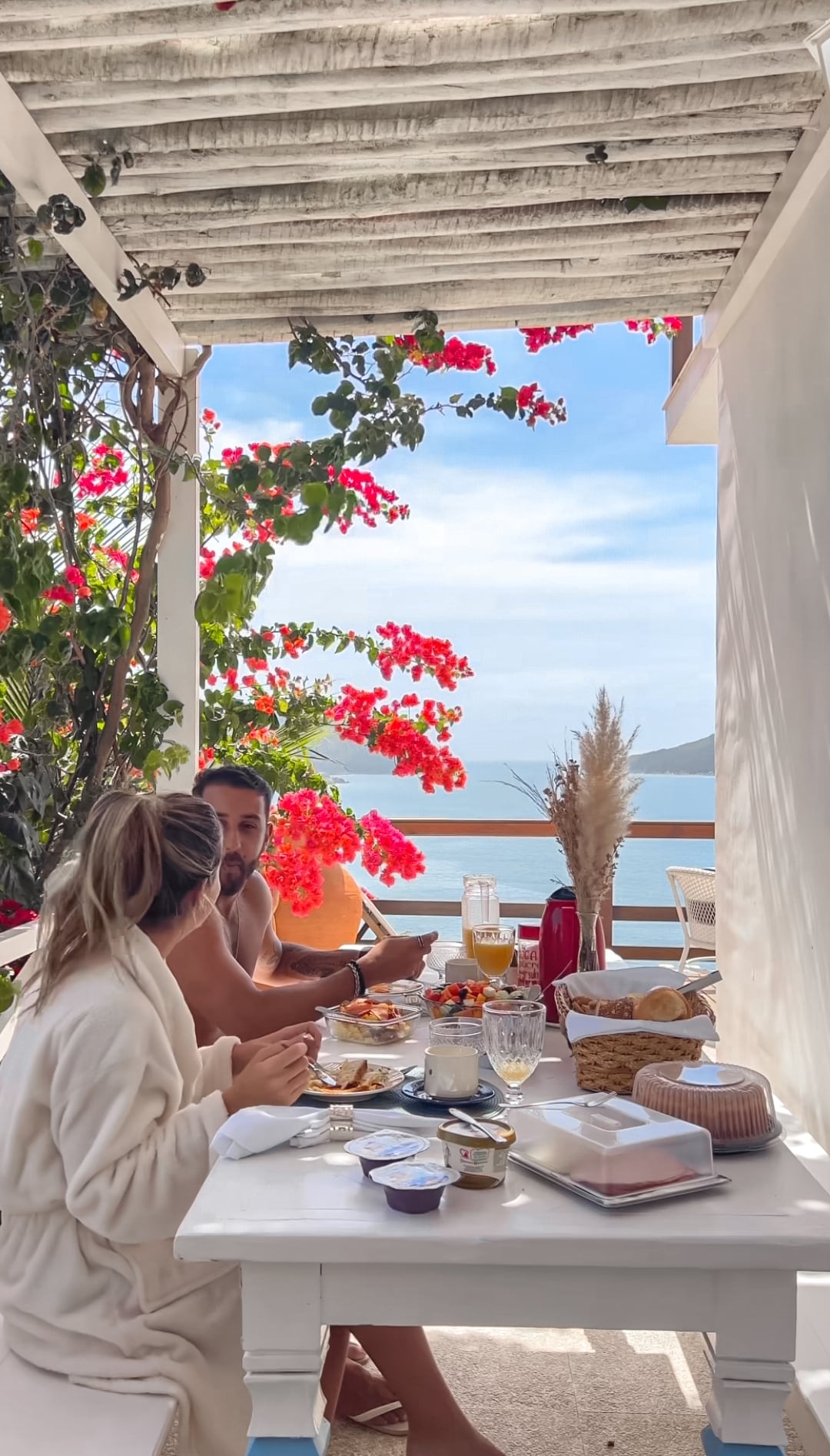 An outdoor dining area is adorned with vibrant bougainvillea, providing a scenic backdrop of the ocean. A white table is set with breakfast items, creating a welcoming atmosphere for enjoying meals al fresco. Light filters through the surrounding foliage, enhancing the serene setting.