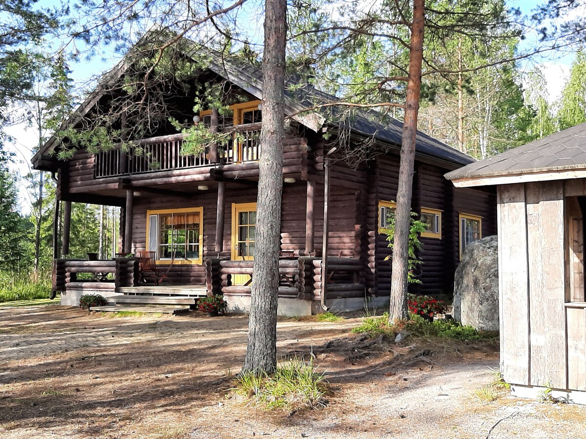 A wooden cabin with a multi-level structure features a welcoming porch and vibrant yellow window frames. The surrounding nature includes tall trees and a well-maintained yard, enhancing the cabin's rustic charm. An additional smaller building is positioned nearby.
