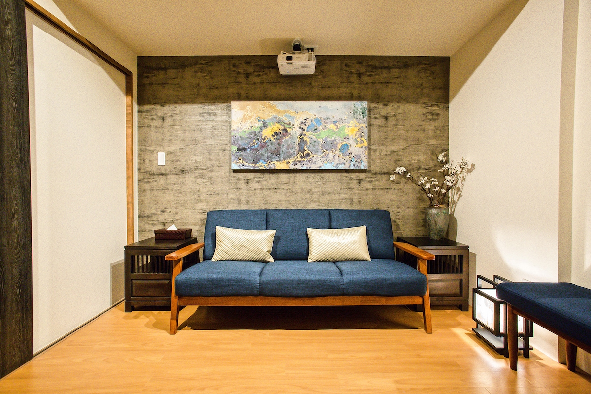 A cozy living area is highlighted by a blue sofa with decorative cushions. A piece of abstract art is mounted on the textured wall above, and small side tables are positioned on either side of the sofa, complementing the wooden floor.