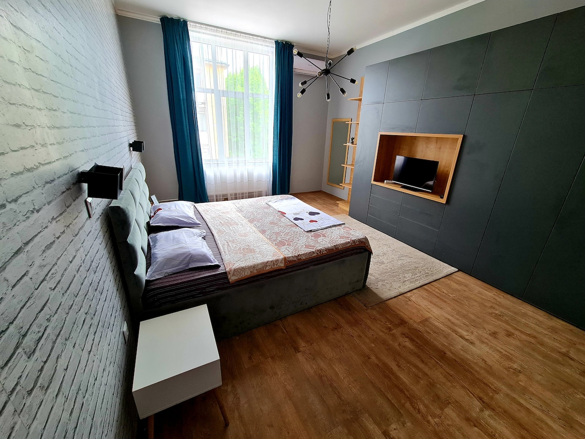 A spacious bedroom features a comfortable bed with a decorative blanket and soft pillows. A sleek nightstand is positioned beside the bed. A large window draped with blue curtains allows natural light to fill the room, while a modern entertainment unit is integrated into the wall.