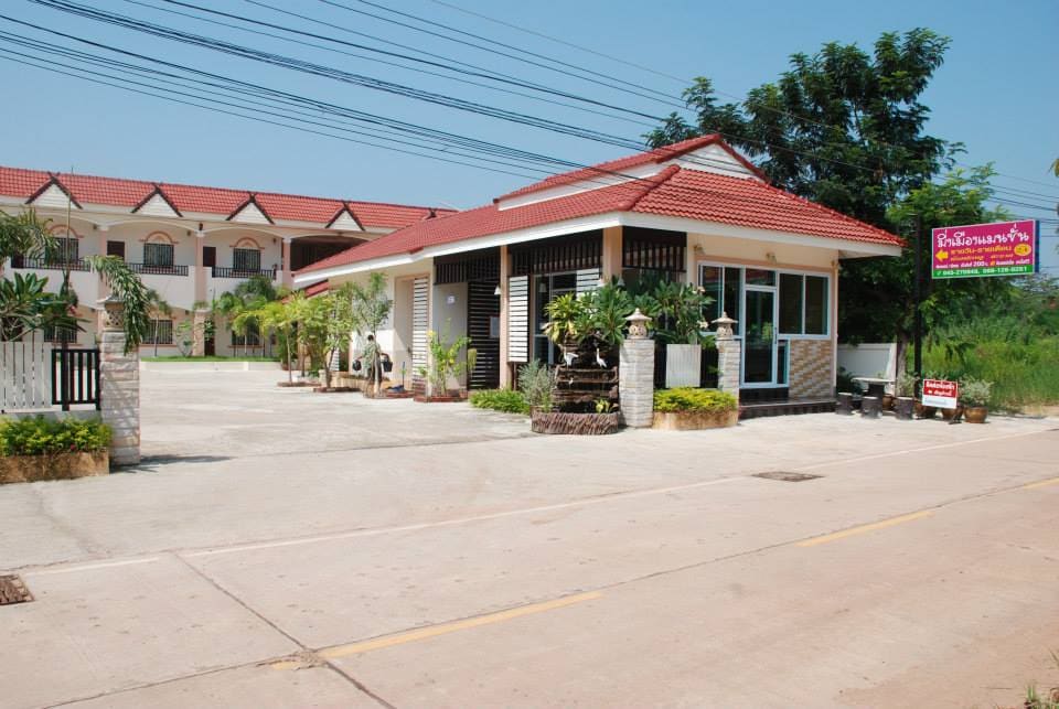 The entrance area features a well-maintained façade with a red-tiled roof and large windows. A spacious parking lot is visible, surrounded by landscaped greenery. The surrounding buildings echo a similar architectural style, set against a clear blue sky.