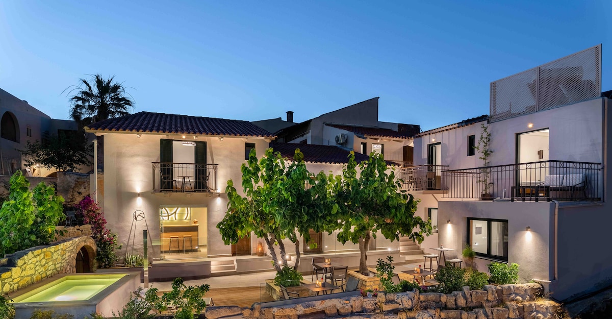 The exterior of Kamares Sanctuaries showcases a charming complex of renovated villas and suites, accented by lush greenery and stone pathways. Warm, soft lighting enhances the inviting atmosphere as twilight sets in. A shared outdoor area features seating arrangements and a glimpse of the tranquil surroundings.