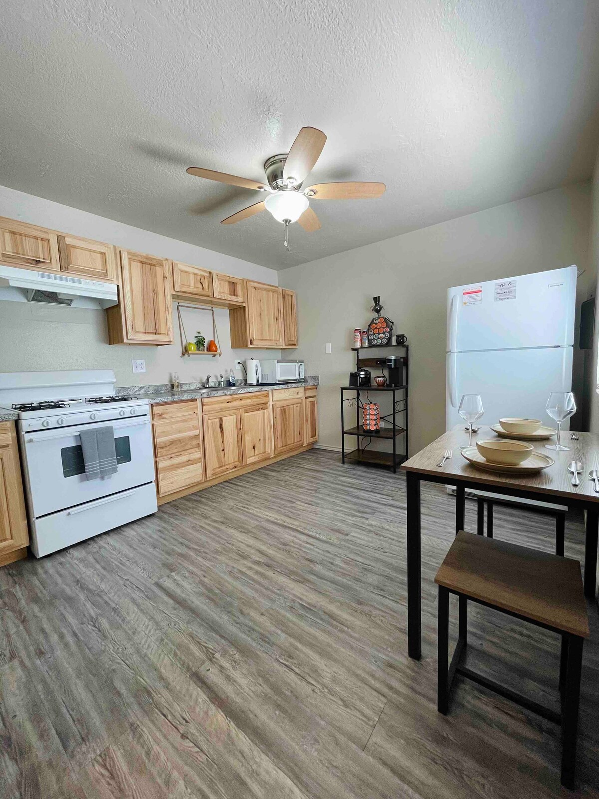 The kitchen features natural wood cabinetry and a ceiling fan, providing a functional workspace. Essential appliances include a stove, oven, microwave, and refrigerator. A small dining table with two chairs is present, alongside glassware and dishware, creating an inviting area for meals.
