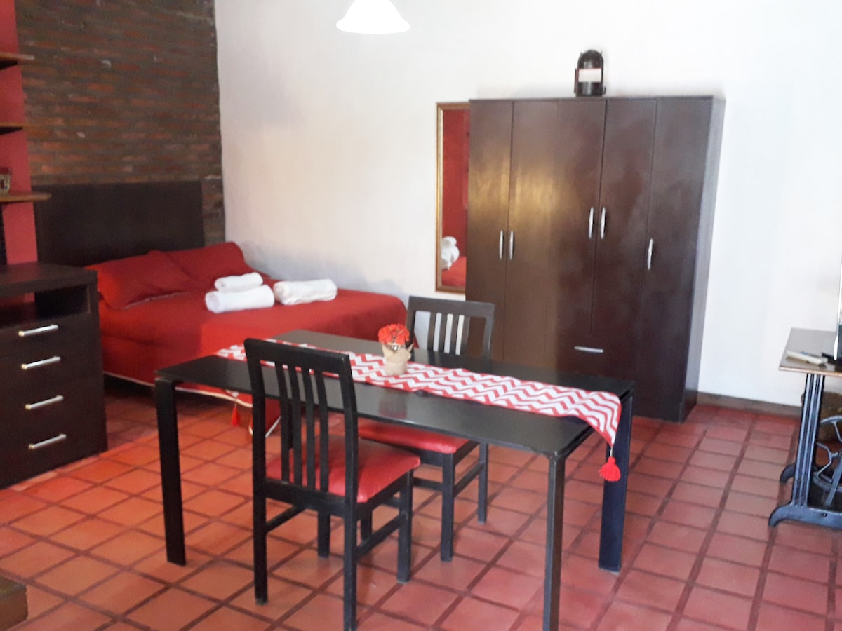 An inviting living space is depicted, featuring a red bed with folded towels and a dark wooden dining table with matching chairs. A decorative table runner adds a pop of color, while light from a ceiling fixture illuminates the brick wall background.