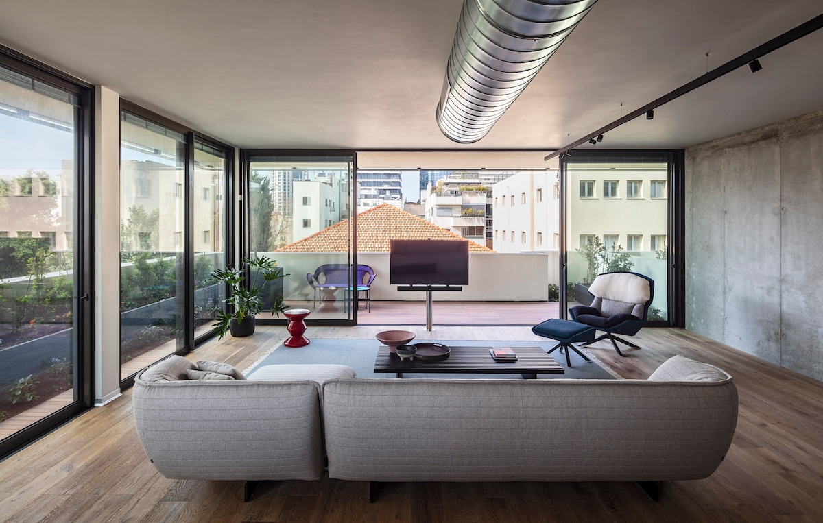 A bright living room features large floor-to-ceiling windows allowing ample natural light. Modern furnishings, including a sectional sofa and accent chairs, are arranged to create an inviting atmosphere. A television is positioned for viewing, while a small outdoor balcony can be seen through the glass.