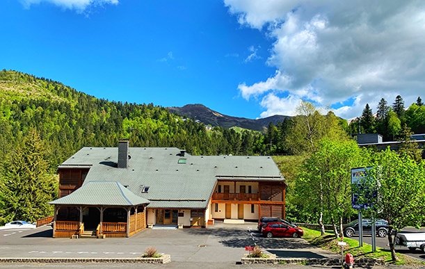 The exterior of a two-story chalet-style building is visible, featuring a gray roof and wooden accents. Surrounding lush greenery and rolling hills create a natural setting. A parking area is shown in the foreground, with cars parked and a sign indicating nearby amenities.