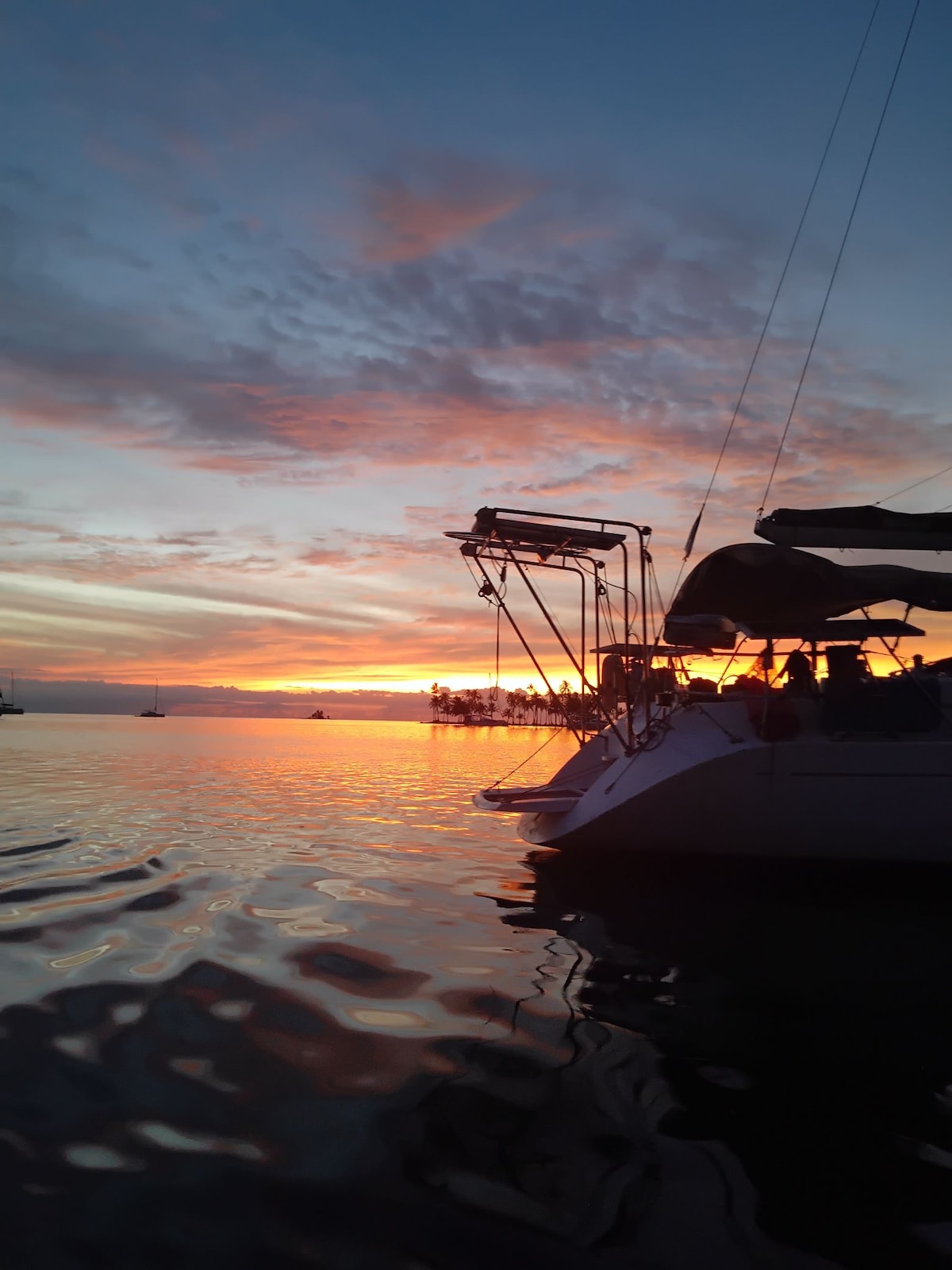 A sailboat is anchored in calm waters, reflecting the vibrant colors of the sunset. The sky is filled with soft hues of orange, pink, and purple, while silhouettes of distant boats and palm trees can be seen against the horizon.