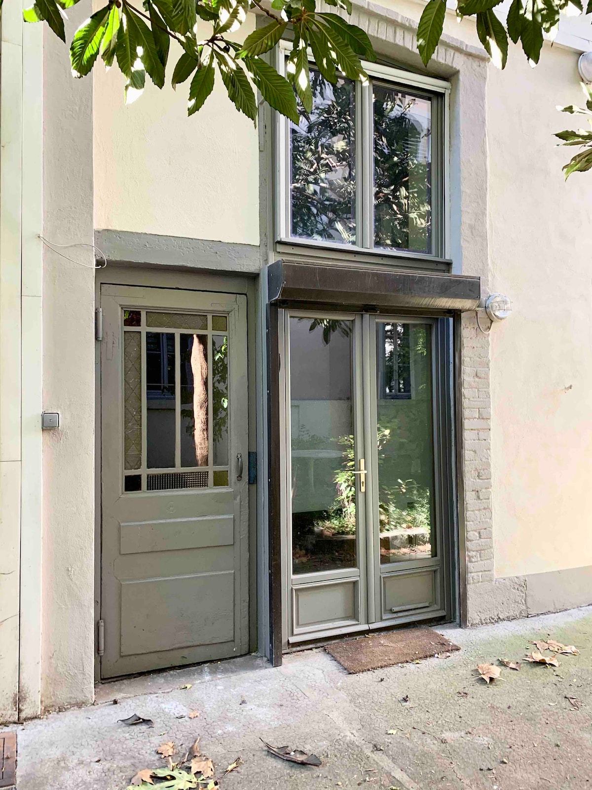 The exterior of the duplex showcases a charming entrance with a large glass door and a smaller, framed door for added access. Natural light filters through the windows, creating a welcoming atmosphere. Lush greenery is visible, enhancing the tranquil setting.