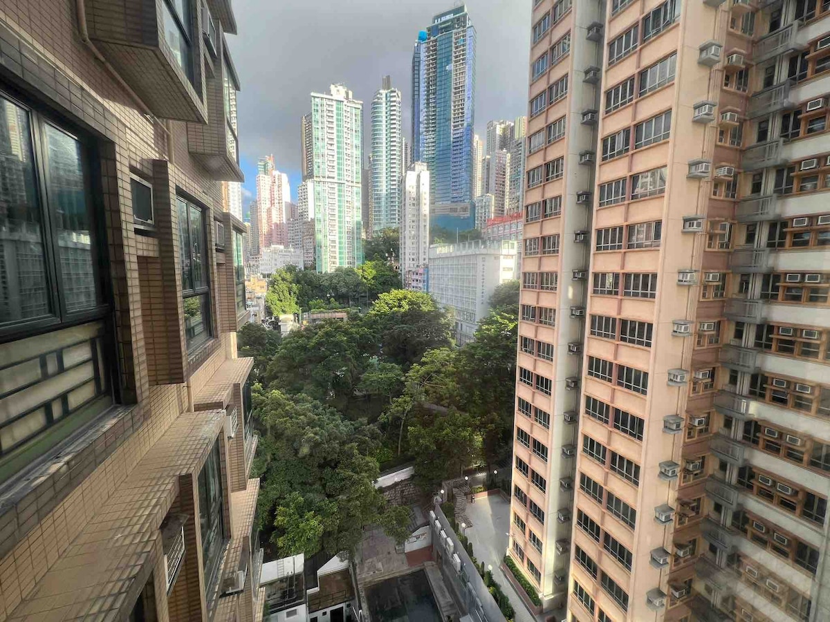 A view of the cityscape is displayed, showcasing a mix of modern skyscrapers and lush greenery. Tall buildings stand against a backdrop of cloudy skies, while trees can be seen filling the space below, providing a contrast to the urban environment.