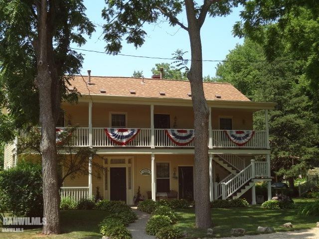 A charming two-story building features a covered porch with decorative railings. The exterior is painted in a soft hue, complemented by large windows. Bunting in patriotic colors adorns the balconies, while trees provide shade around the entrance, enhancing the inviting facade.