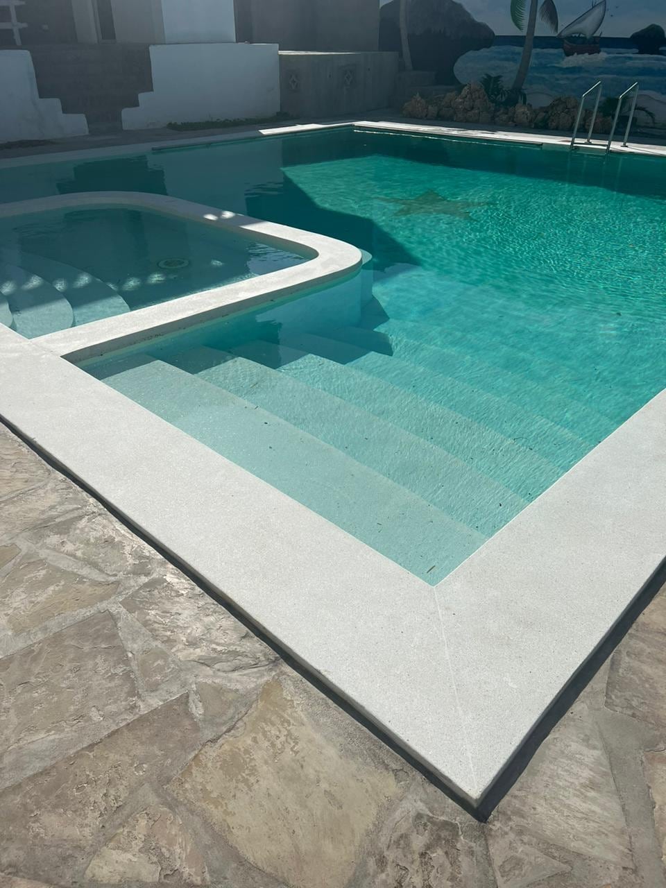 A clear swimming pool with a separate hot tub is surrounded by a smooth stone deck. Steps lead gently into the water, highlighting the inviting turquoise hue. The area is well-lit, enhancing the tranquil ambiance of the space.