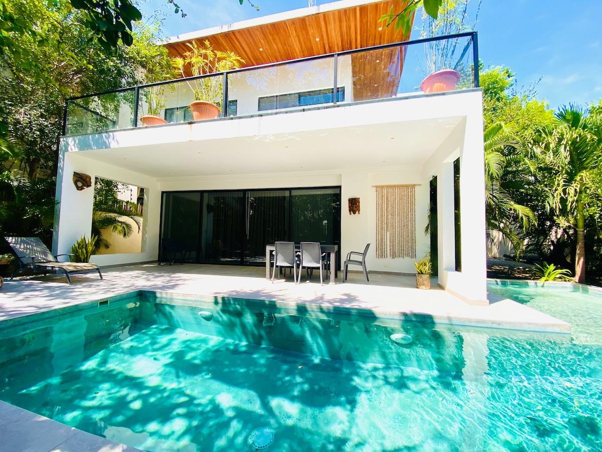 A spacious modern villa is showcased with a deep swimming pool reflecting the clear sky. The ground floor features a patio with outdoor seating, while large glass doors provide a seamless view of the pool area surrounded by lush greenery.