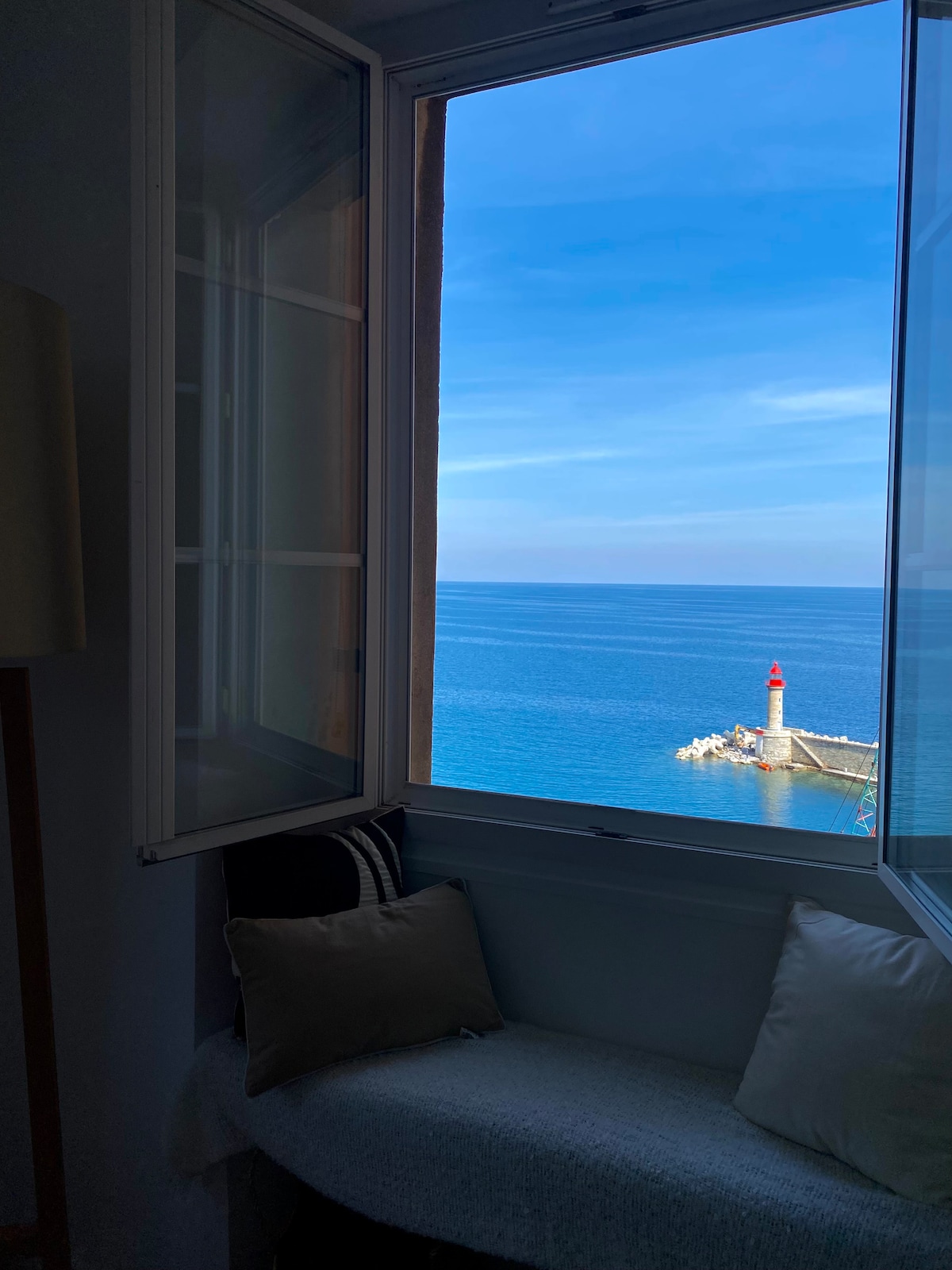 A view from an open window reveals calm blue waters and a distant red and white lighthouse at the harbor entrance. The light filtering through the window illuminates a cozy seating area with cushions, enhancing the serene coastal ambiance.