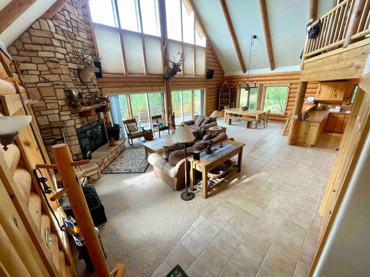 A spacious living area is filled with natural light from large windows that extend to the peak of the vaulted ceiling. A stone fireplace serves as a central feature, complemented by comfortable seating arrangements and a dining table. The open layout creates a seamless flow to the kitchen area.