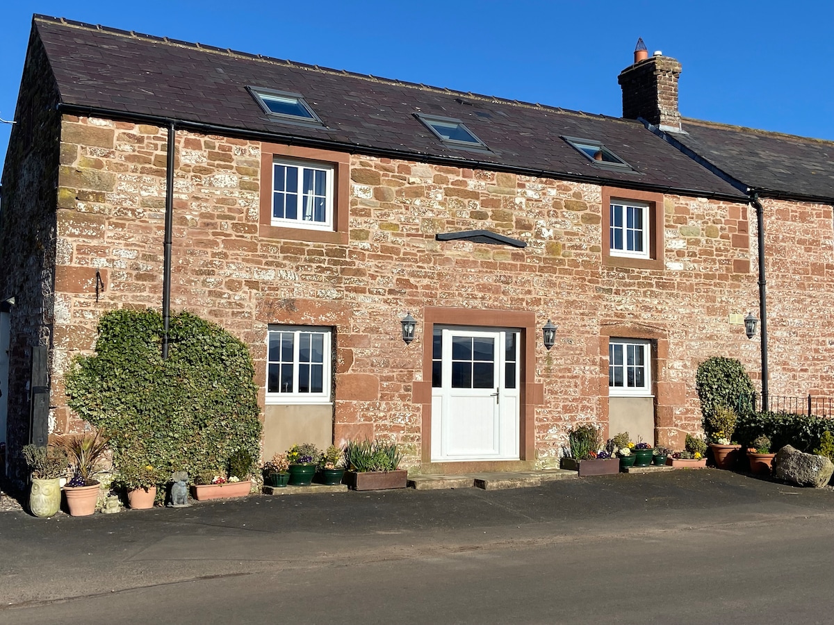 A charming barn conversion is featured, showcasing a stone exterior with multiple windows on the upper level. Flower pots line the front, adding a touch of greenery. A welcoming entrance with double doors is centrally positioned, inviting guests into the cozy accommodation.