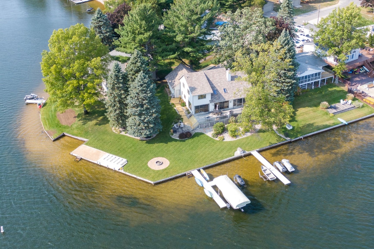 An aerial view shows a spacious lakefront property surrounded by trees. Two docks extend into the water, and a sandy shoreline leads up to a well-maintained house. A fire pit is visible in the grassy area near the shore, offering a relaxing outdoor space.