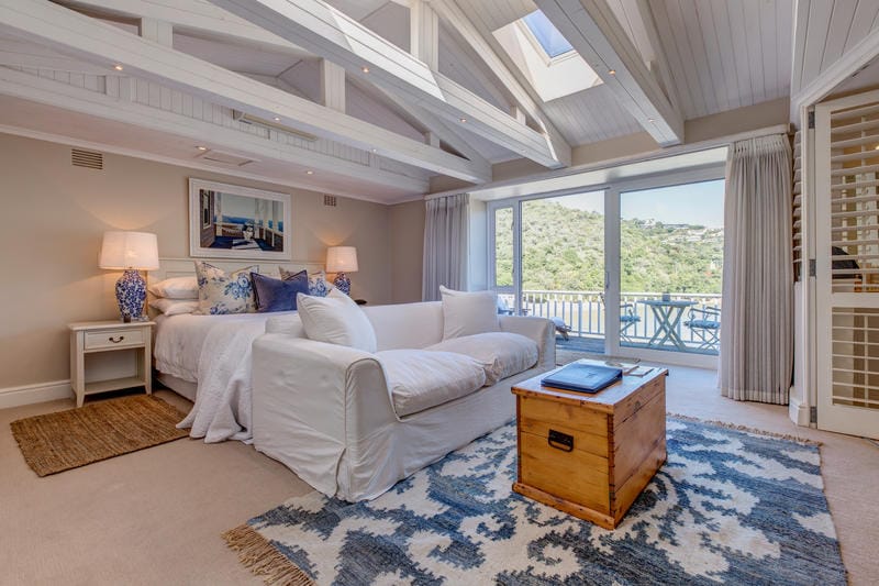 A spacious room features a large bed with decorative pillows, accompanied by a comfortable seating area. Natural light floods in through the large glass doors, providing views of the lagoon and surrounding nature. Soft textures and neutral tones create a serene environment.