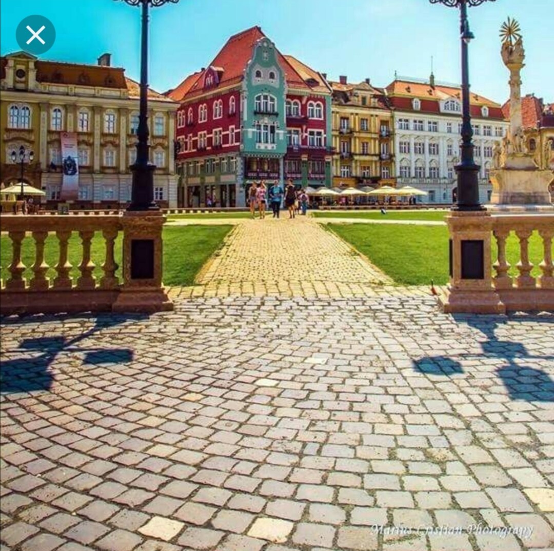 A vibrant square is viewed from a cobblestone path, flanked by ornamental lamp posts. Historic buildings in a variety of colors line the perimeter, with people strolling leisurely. Lush green grass and outdoor seating areas contribute to a lively and inviting atmosphere.
