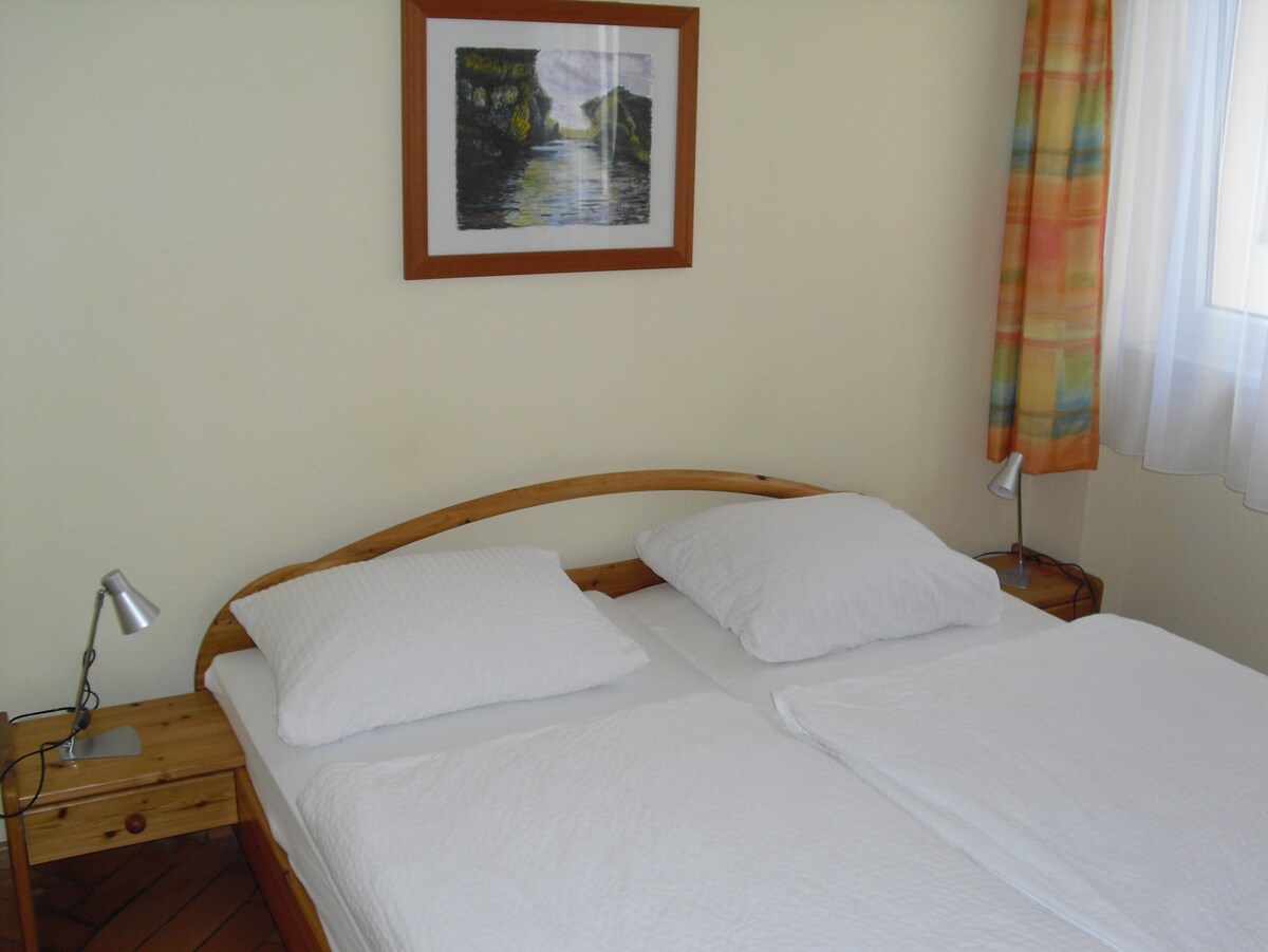 A simple, modern bedroom features a spacious bed with two white pillows and blankets. A wooden headboard adds a natural touch. Two bedside lamps are positioned on either side. A framed artwork adorns the wall, while light-colored curtains gently diffuse the daylight streaming through the window.