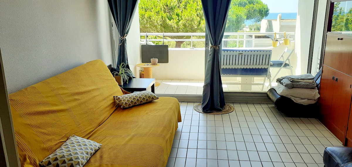 A spacious living area is visible, featuring a yellow sofa bed along with decorative pillows. Natural light enters through the sliding glass doors, leading to a balcony with a sea view. The tiled floor adds a clean and open feel to the room.