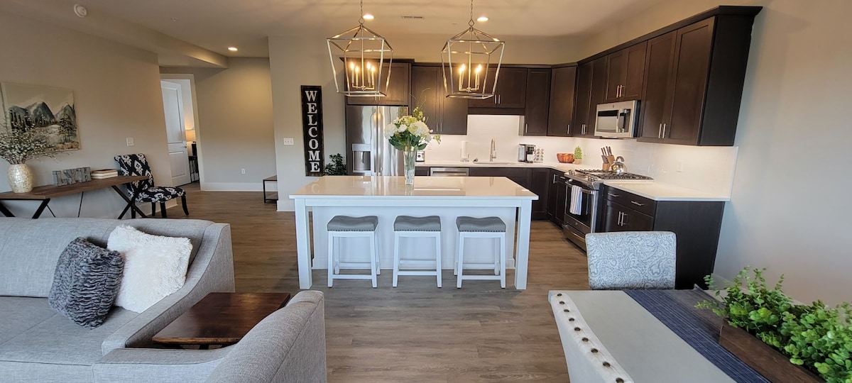The modern kitchen features dark cabinetry and stainless steel appliances, with a large island at its center. Three high-backed stools are arranged around the island. A cozy seating area, with a light grey sofa and decorative cushions, is positioned nearby.