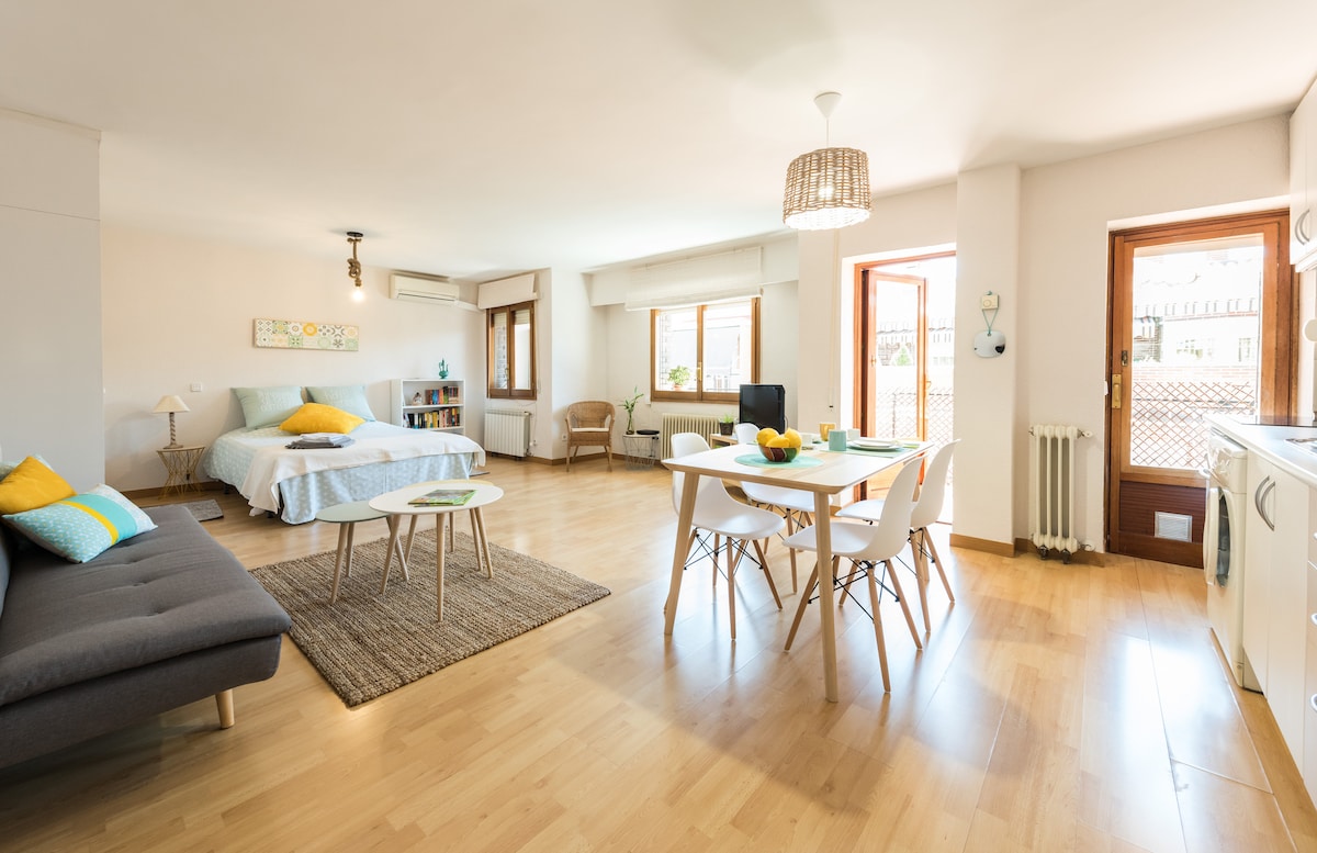 A well-lit studio space features a spacious bed with light bedding, a cozy seating area with a gray sofa, and a dining table for four. Large windows allow natural light to fill the room, enhancing the welcoming atmosphere, while a wooden floor adds warmth.