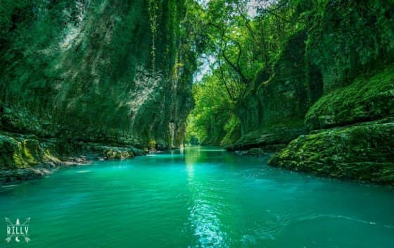 A serene river flows through a lush canyon, flanked by towering green cliffs. Sunlight filters through the dense foliage, casting dappled reflections onto the water's surface. The vibrant turquoise hue of the water contrasts beautifully with the surrounding greenery.
