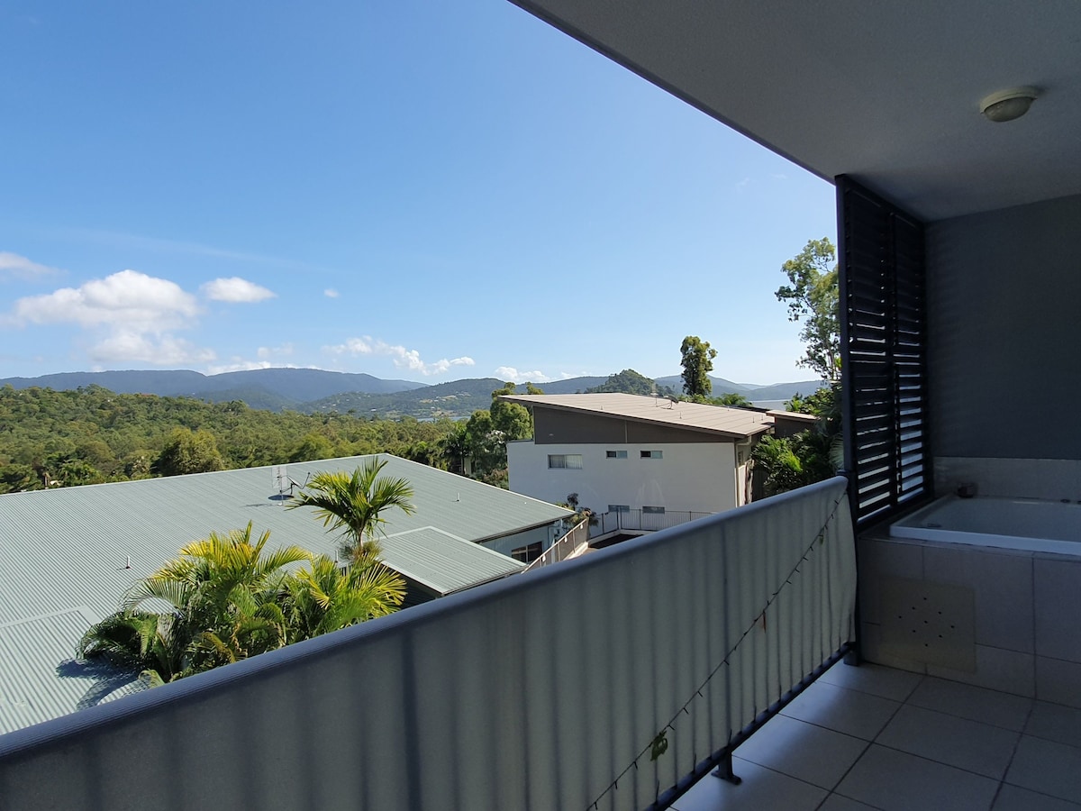 A balcony view showcases lush tropical greenery and distant mountain ranges under a clear blue sky. The railing frames the scene, while a portion of the private spa is visible in the corner, inviting relaxation in a serene environment.