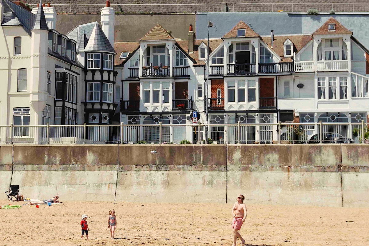 The seafront view showcases a row of heritage-style houses with distinct architectural features. Sand stretches in the foreground, where a few children are playing. The sea wall and gentle waves create a serene coastal ambiance, highlighting the proximity to the beach.