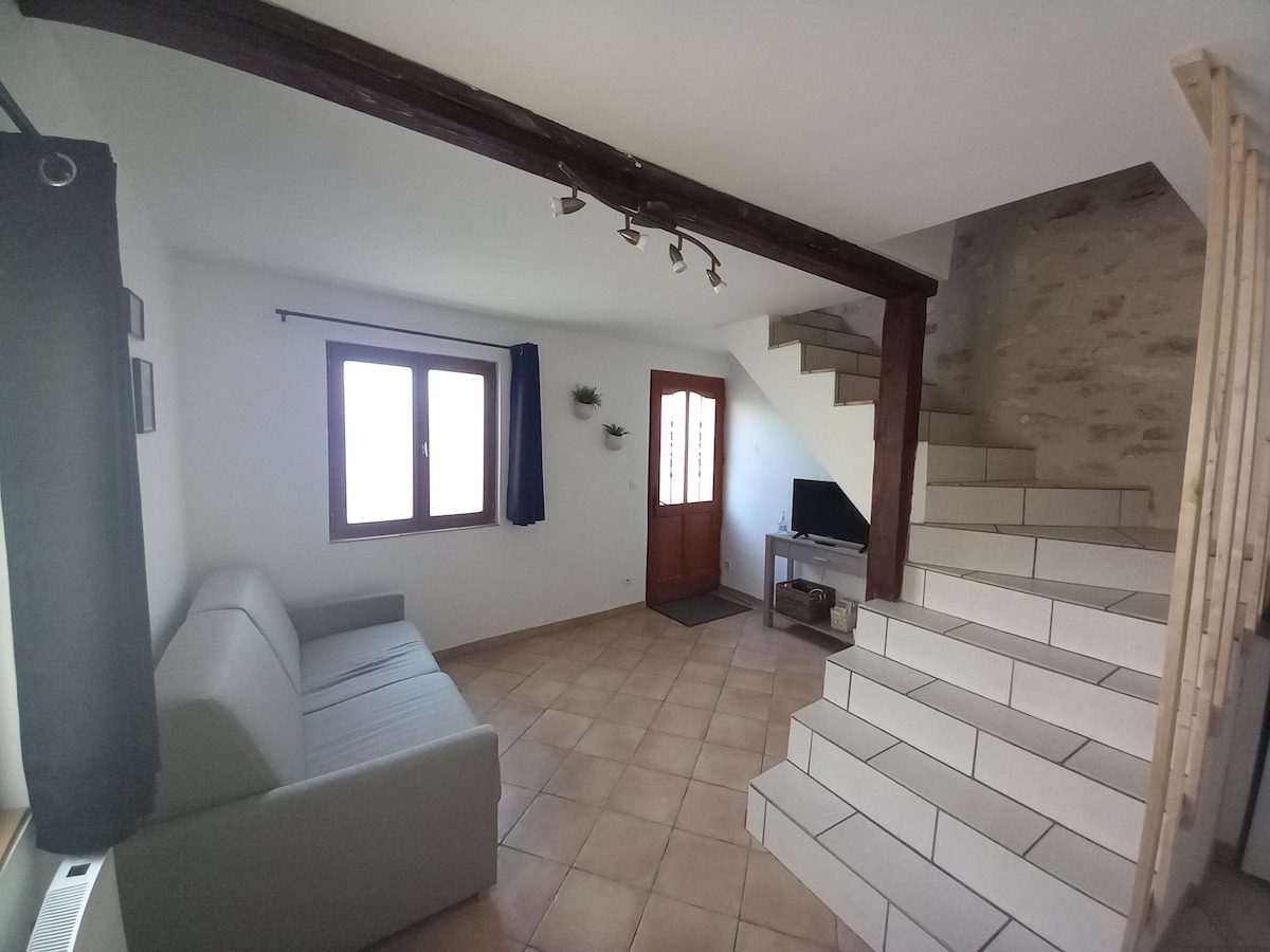 The living area features a comfortable gray sofa and two windows allowing natural light to fill the space. A wooden staircase leads to the upper level, while a decorative wooden beam spans the ceiling. A television stands in the corner, complemented by a welcoming entrance door.