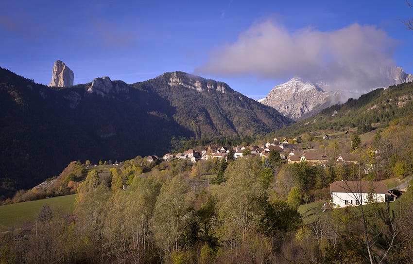 The image captures a serene landscape featuring rolling hills and a picturesque village nestled among trees. Rocky mountains rise in the background, partially shrouded in clouds, while a peaceful atmosphere is conveyed by the lush greenery and distant peaks.