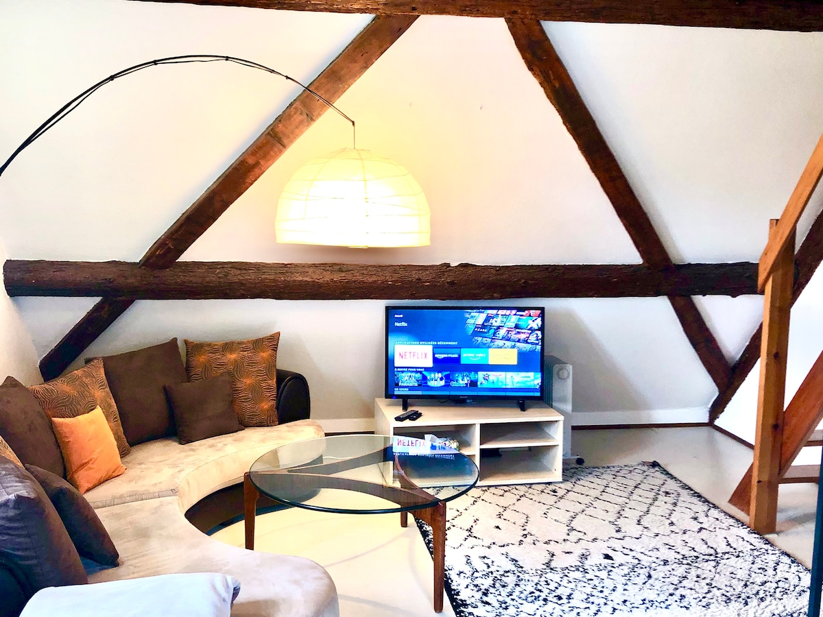 A cozy living area features a curved sofa adorned with cushions, a glass coffee table, and an area rug. A wall-mounted TV offers various entertainment options. Exposed wooden beams add warmth to the space, with gentle lighting enhancing the room's ambiance.