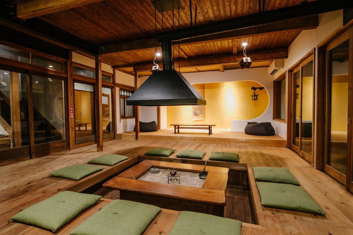 A spacious dining area features a central sunken hearth surrounded by green floor cushions. Rustic wooden beams enhance the warm atmosphere. Large glass doors provide a view of the outdoors, while softly lit walls create an inviting environment for shared meals.