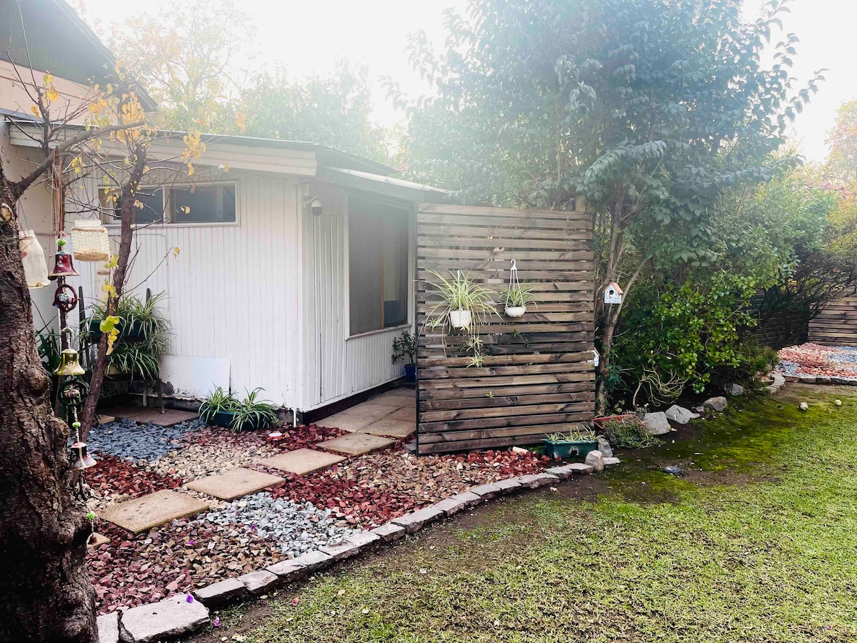 An exterior view of the guest house is presented, featuring a wooden privacy screen and a stone pathway leading to the entrance. Lush greenery and decorative plants are visible along the garden, creating a serene outdoor environment. A small grassy area enhances the tranquility of the space.